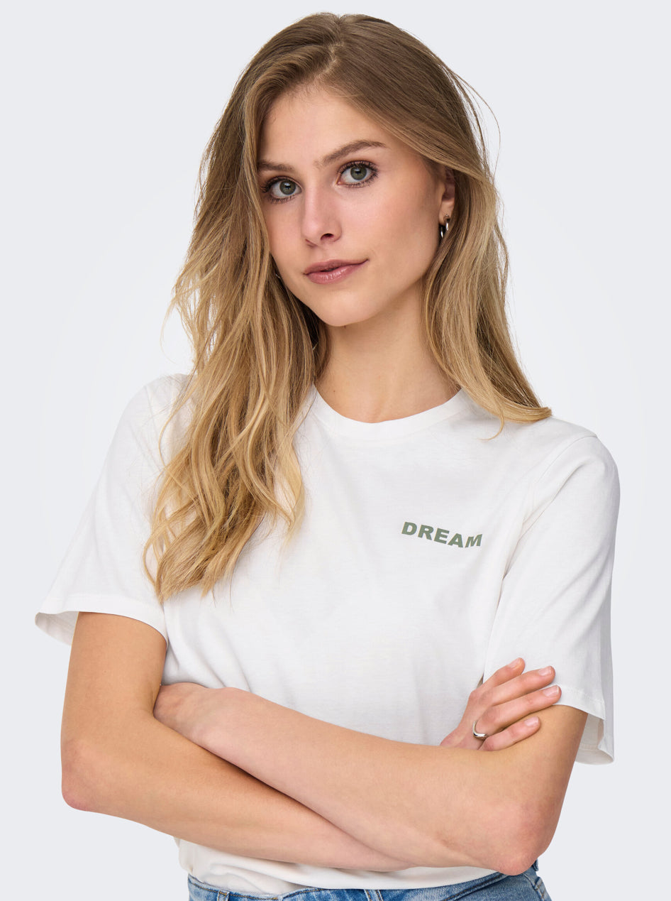 Hvid tee fra JDY. Ed lille print foran hvor der står Dream , på ryggen er der et stort print med blomster, t-shirten ses her på model der står med fronten til.